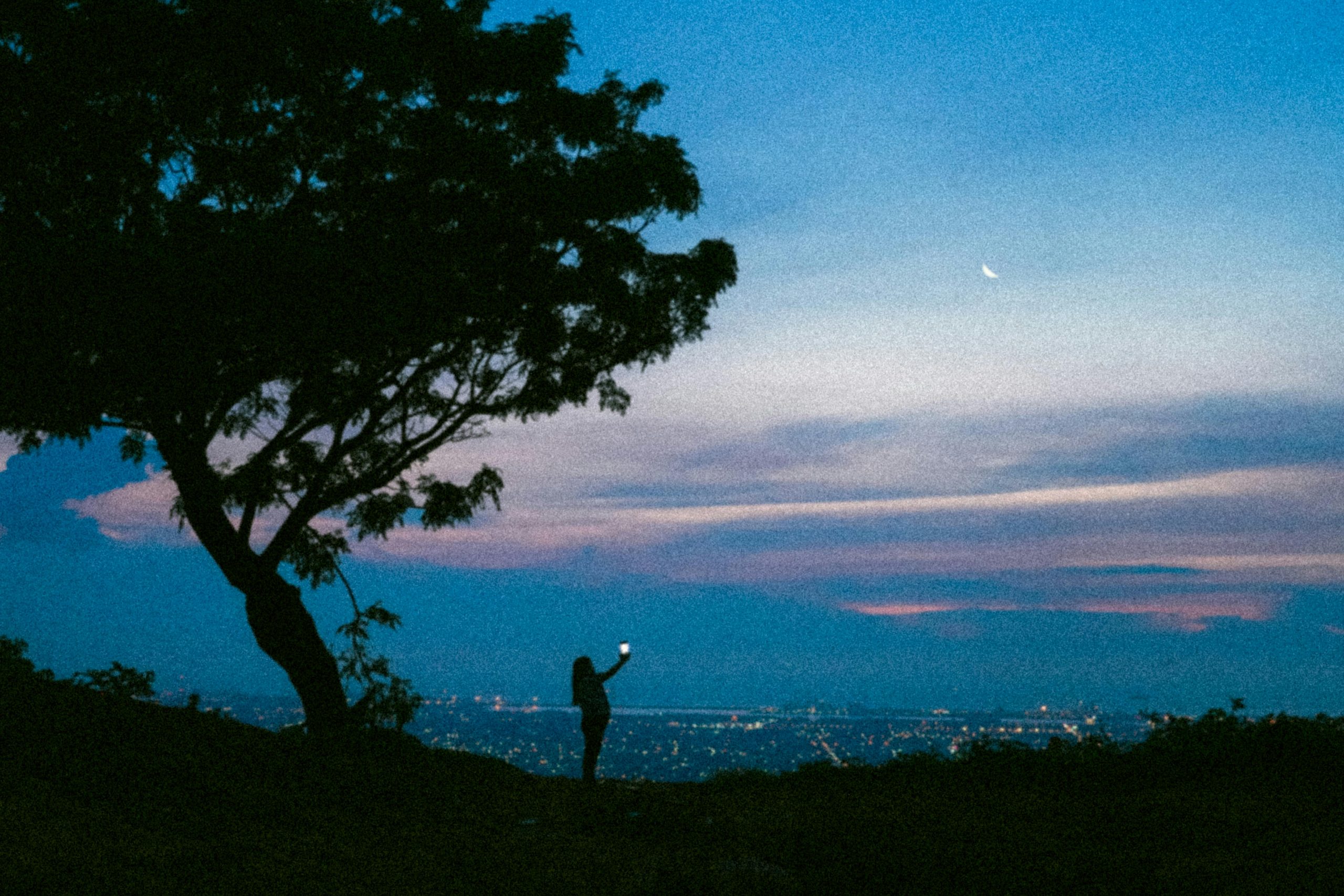 A silhouette of a person taking a photo against a cityscape during dusk with a visible crescent moon.