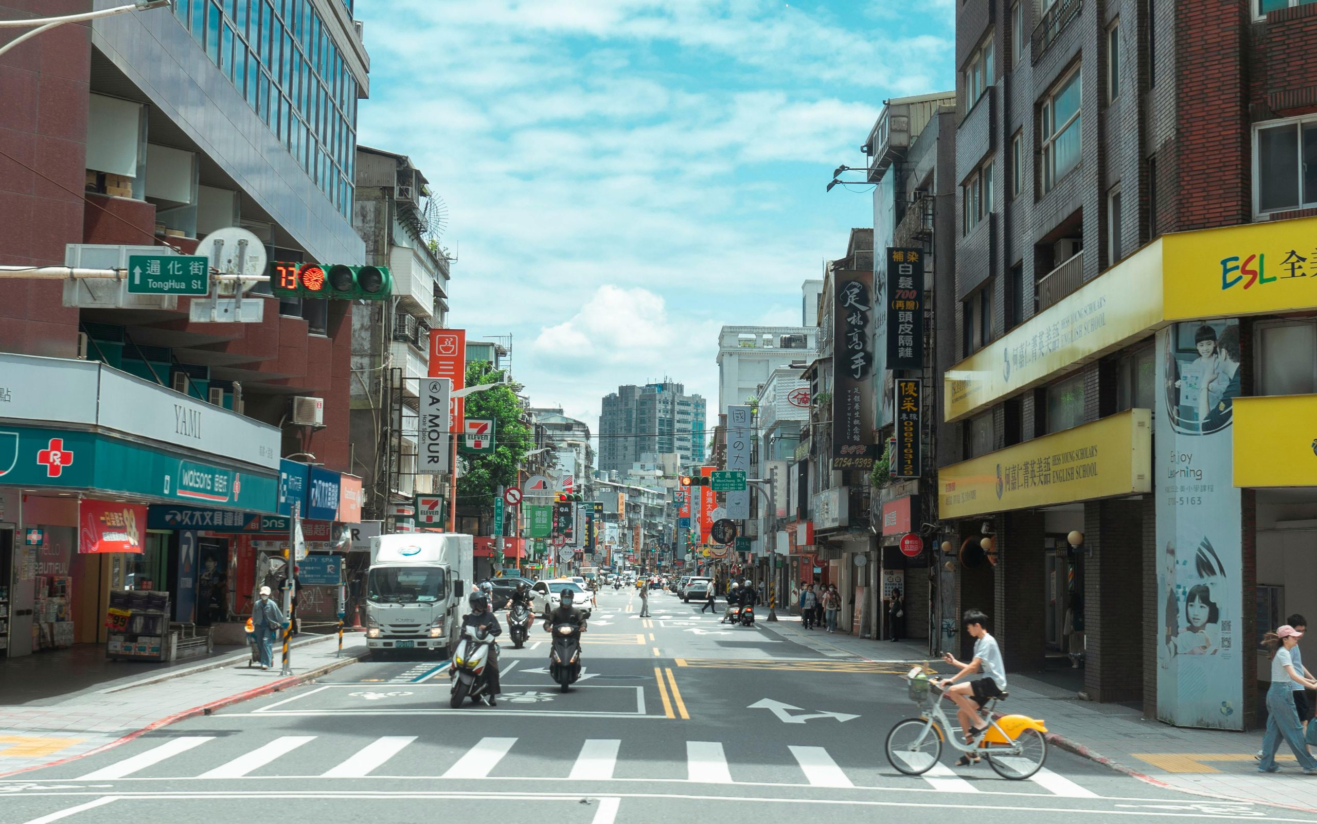 A bustling Taipei street with people, cars, and scooters under a bright sky in Taiwan.